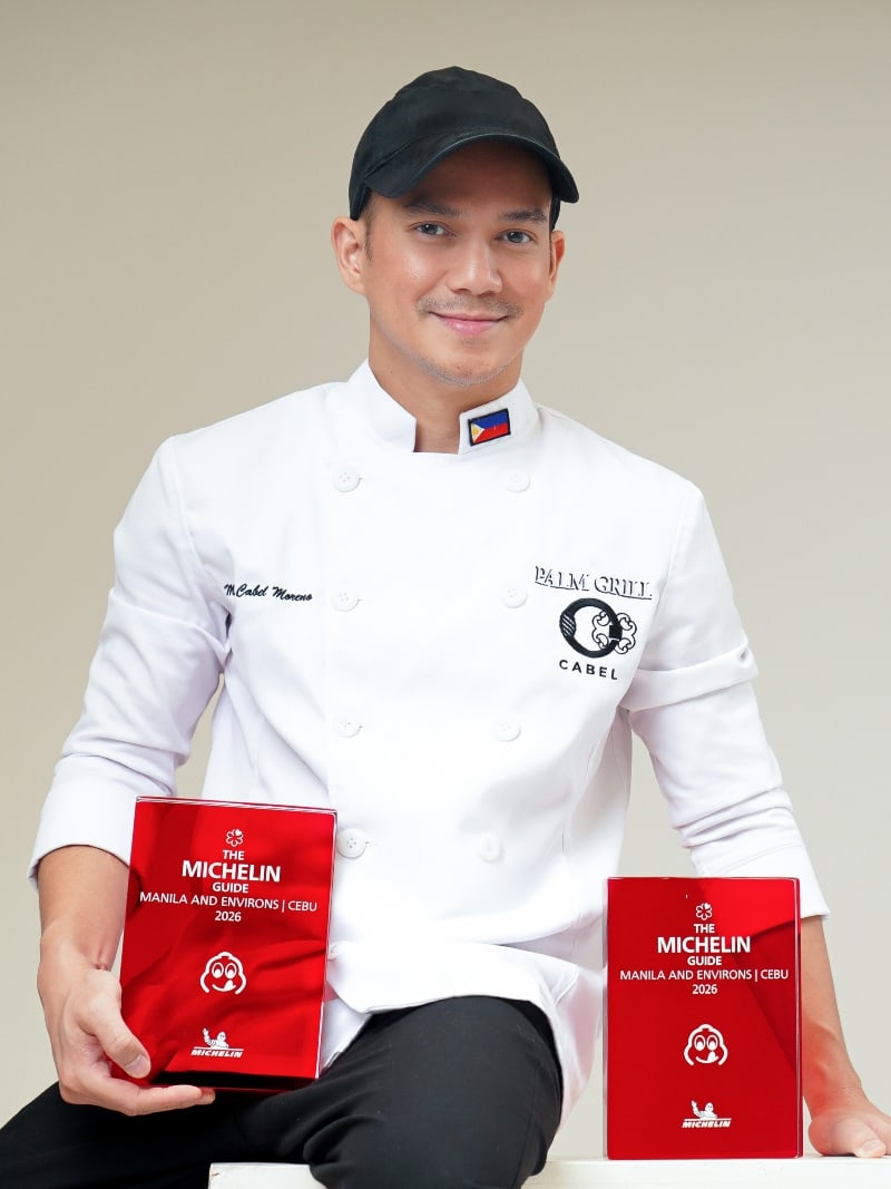 Chef Miggy Cabel Moreno in his chef’s coat and cap, holding his two MICHELIN Guide 2026 plaques